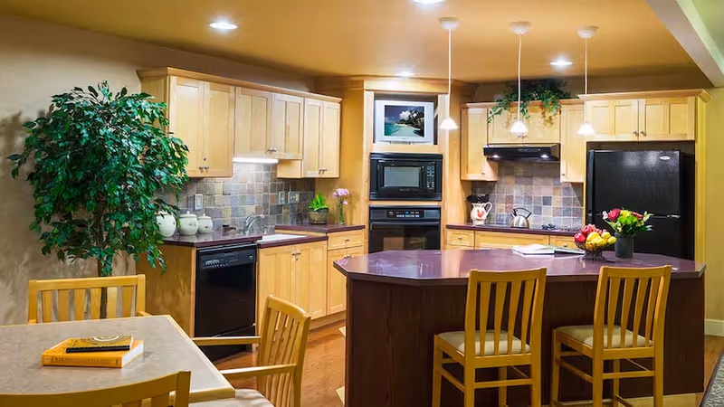 Bright kitchen with light wood cabinets, a central island with three chairs, appliances, pendant lights, and a small dining table to the left.