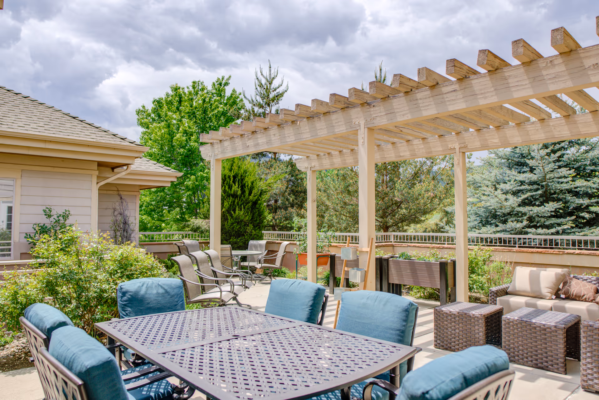 Outdoor patio area at Golden Pond Senior Living featuring a metal table with blue cushioned chairs, a wooden pergola, additional seating with wicker furniture and cushions, surrounded by greenery and trees under a cloudy sky.