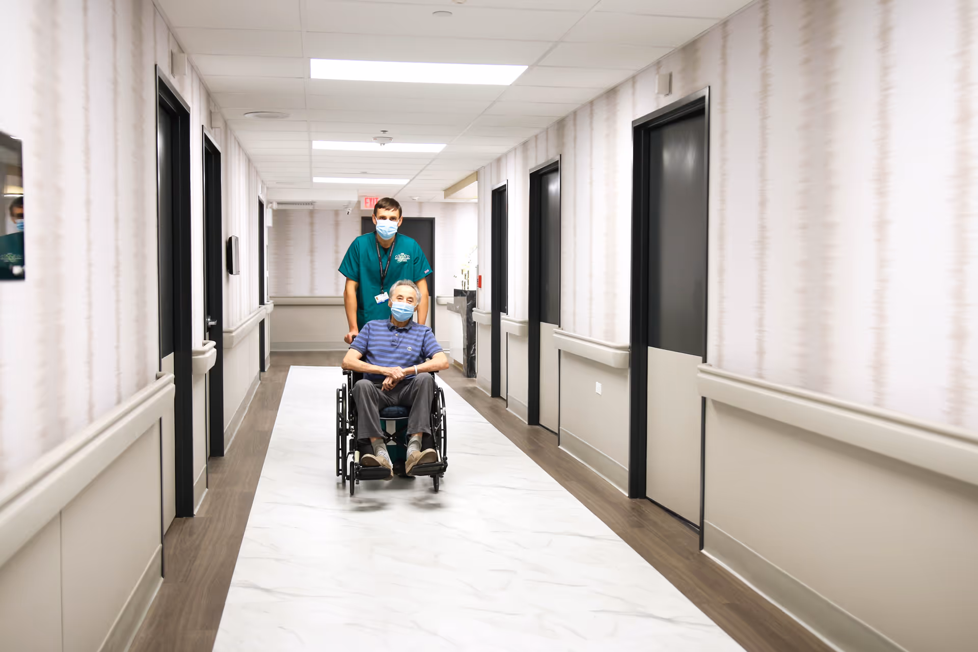A healthcare worker wearing a green uniform and a face mask is pushing an elderly man in a wheelchair down a well-lit hallway with patterned wallpaper and multiple closed doors on either side. Both individuals are wearing face masks.