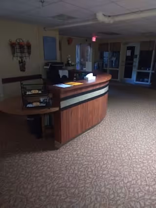 Reception desk area inside a senior living facility with a curved wooden counter, papers and a tissue box on the desk, carpeted floor, and dim lighting. There are doors and windows in the background.
