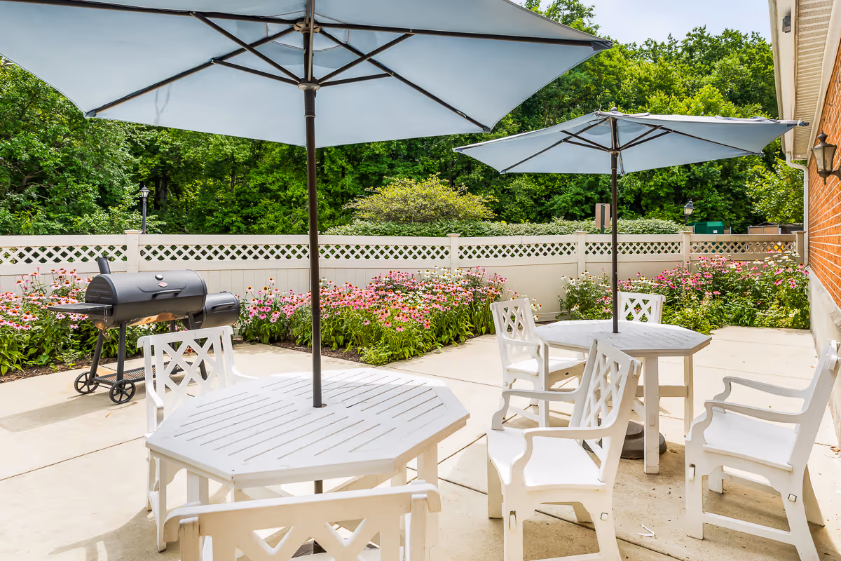 Sunlit patio with white tables and chairs under blue umbrellas, a grill, and flower beds along a lattice fence.