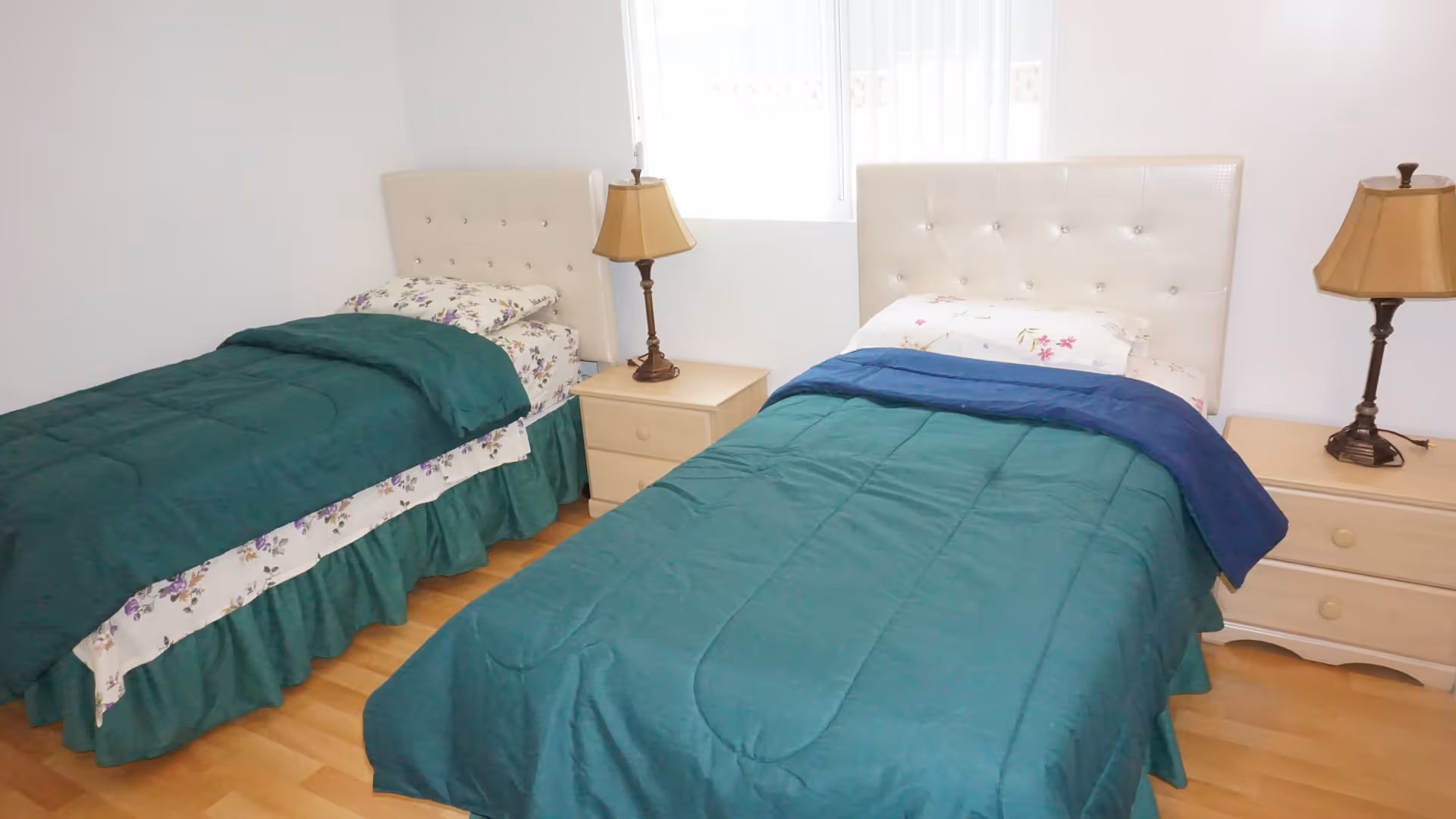 A bedroom with two single beds, each with a green comforter and floral sheets. Between the beds are two wooden nightstands, each with a lamp. A window with vertical blinds is visible behind the beds.