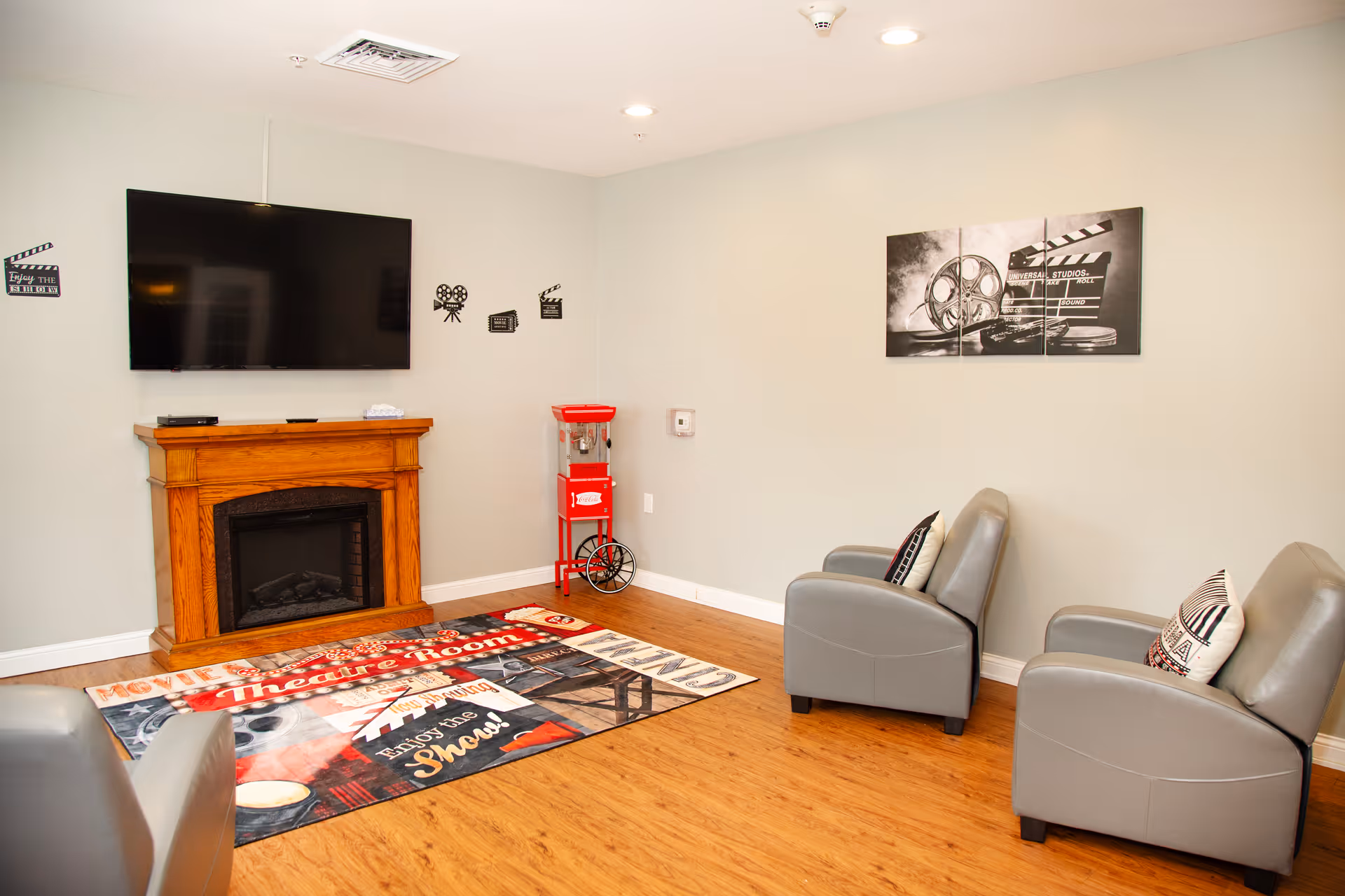 A cozy living room with a wall-mounted flat-screen TV above a wooden electric fireplace. The room features three gray armchairs with decorative pillows, a colorful movie-themed rug, and a red popcorn machine in the corner. The walls are decorated with black and white movie-themed artwork and decals.