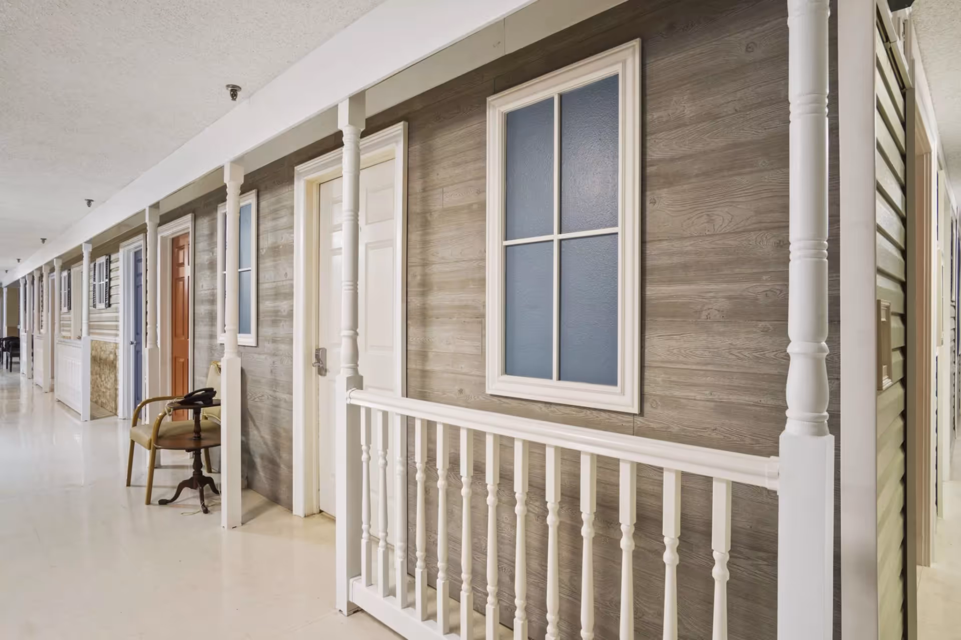 Indoor hallway designed to look like an outdoor porch with multiple doors and windows along a wood-paneled wall, white railings, and a chair with a small table holding a telephone.