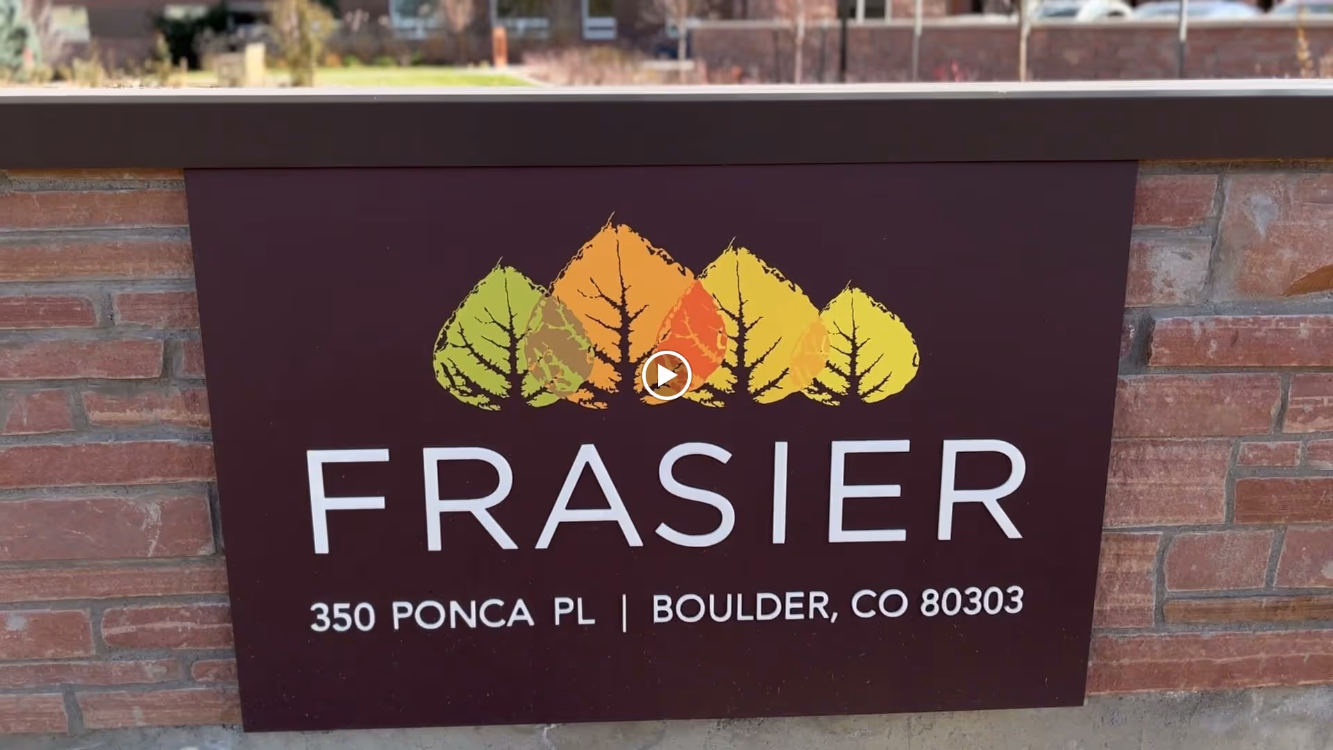 A sign mounted on a brick wall displaying the name 'FRASIER' with colorful leaf graphics above it. Below the name, the address '350 Ponca Pl | Boulder, CO 80303' is written.