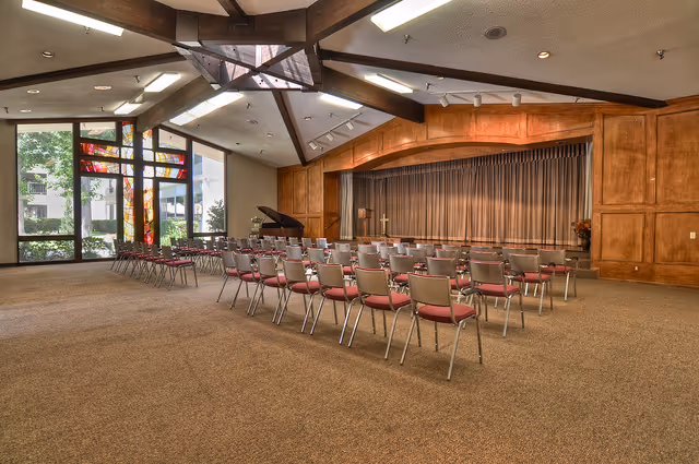 A large room with rows of chairs facing a stage with a closed curtain. The room has a high ceiling with wooden beams and skylights. There is a grand piano near the stage and large windows with colorful stained glass panels on one side, letting in natural light.