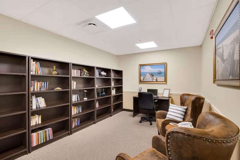 A quiet room with several dark wooden bookshelves partially filled with books and decorative items along one wall. There is a small desk with a black office chair, a laptop, and a framed picture on it. Two brown leather armchairs with a striped pillow on one are positioned near the desk. The walls are light beige with two framed pictures hanging, and the floor is carpeted.