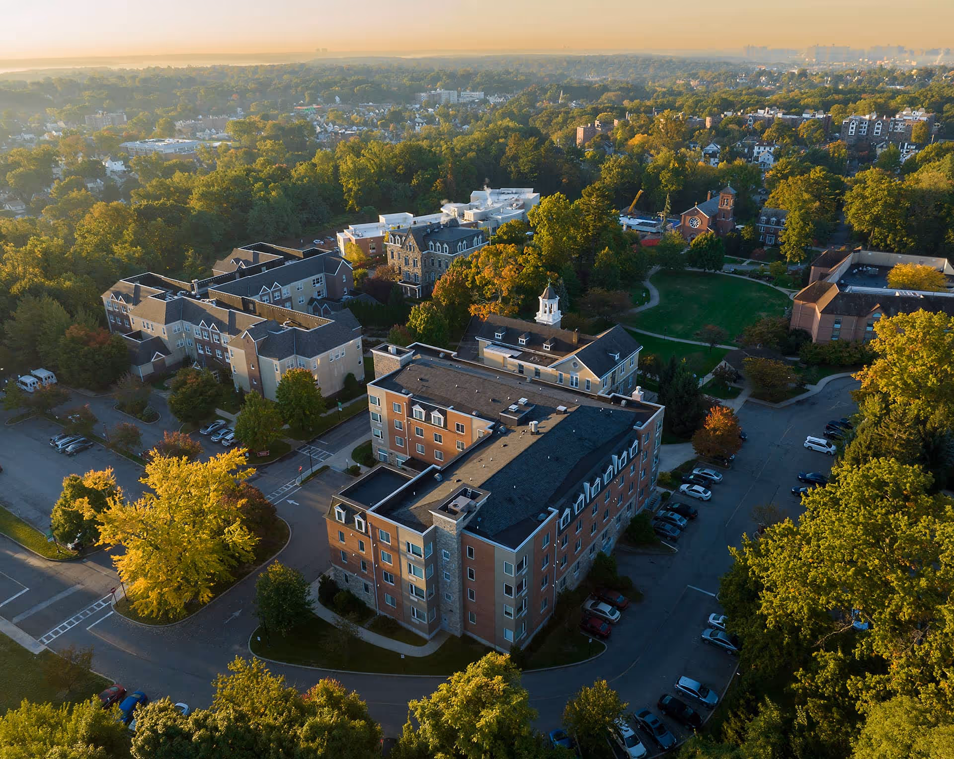 Aerial view of The Wartburg Home facility surrounded by trees and greenery, with multiple buildings, parking lots, and a large grassy area with walking paths, under a clear sky during sunset.