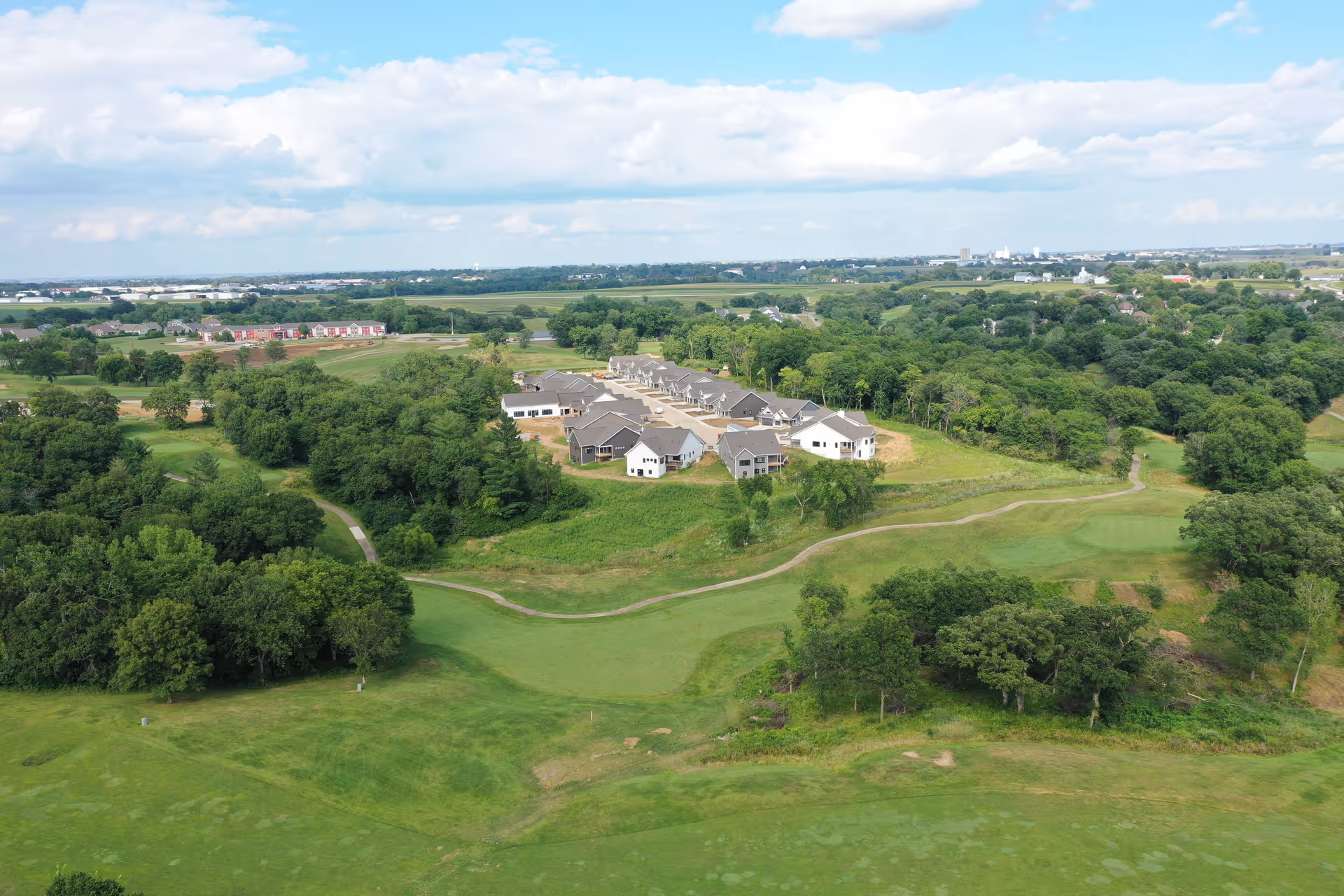 Aerial view of The Estates of Pella: A Vintage Cooperative Community surrounded by green trees and grassy areas, with a winding path leading through the landscape under a partly cloudy sky.