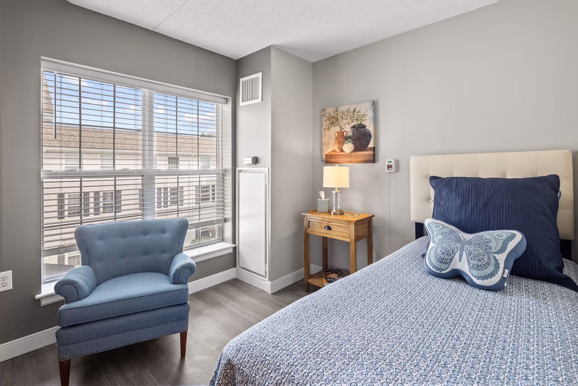 A bright bedroom with a large window featuring white blinds, a blue upholstered armchair, a bed with a blue patterned bedspread and a butterfly-shaped pillow, a wooden nightstand with a lamp, tissue box, and small items, and a wall painting of vases and greenery.