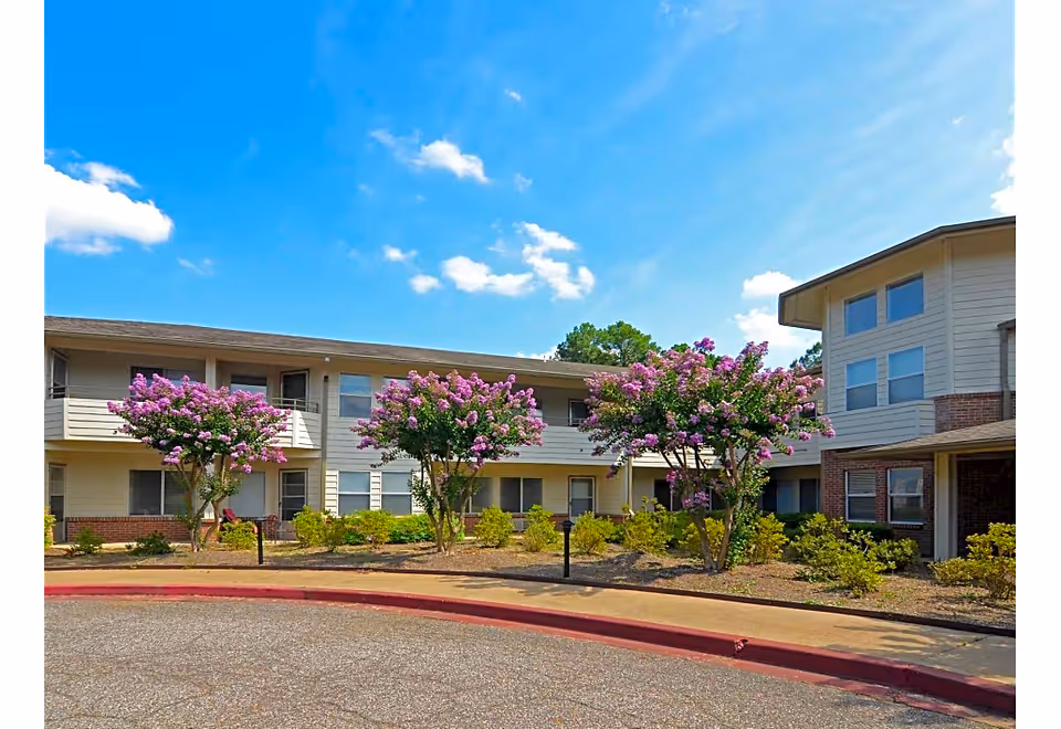 Exterior view of a senior living facility building with two stories, featuring balconies and windows. The foreground shows a curved driveway bordered by a sidewalk and landscaped with flowering trees and shrubs under a bright blue sky with scattered clouds.