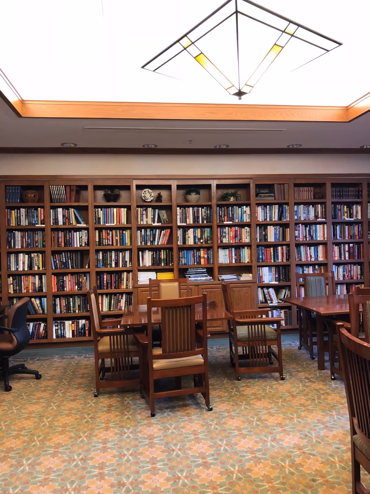 A cozy library/reading room with floor-to-ceiling bookshelves, wooden tables and chairs under a large ceiling light.