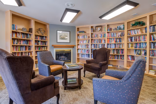 A cozy library room with four upholstered armchairs arranged around a small round wooden table. The walls are lined with built-in wooden bookshelves filled with books and decorative items. A fireplace with a framed picture above it is centered on the far wall. The room has soft lighting from a ceiling fixture.