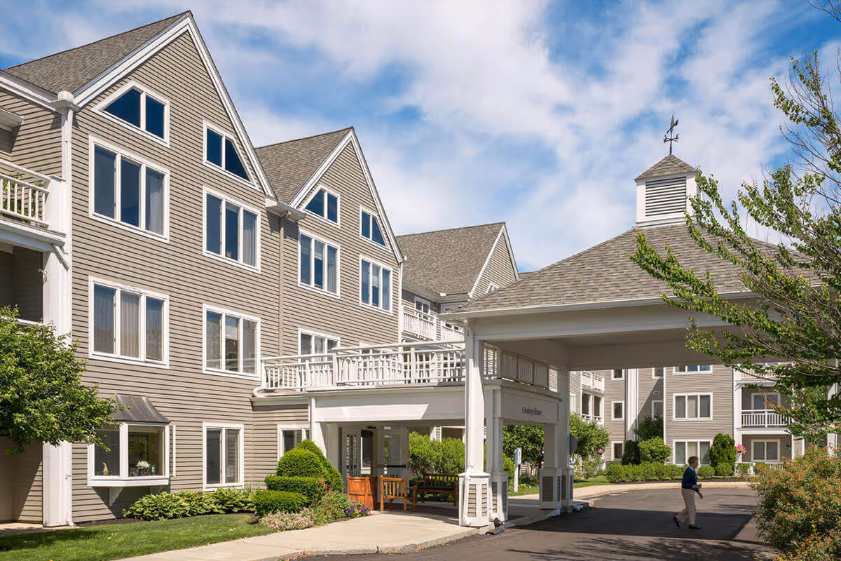 Front exterior of a multi-story senior living building with a covered entrance, balconies, landscaping, and a person walking near the driveway.