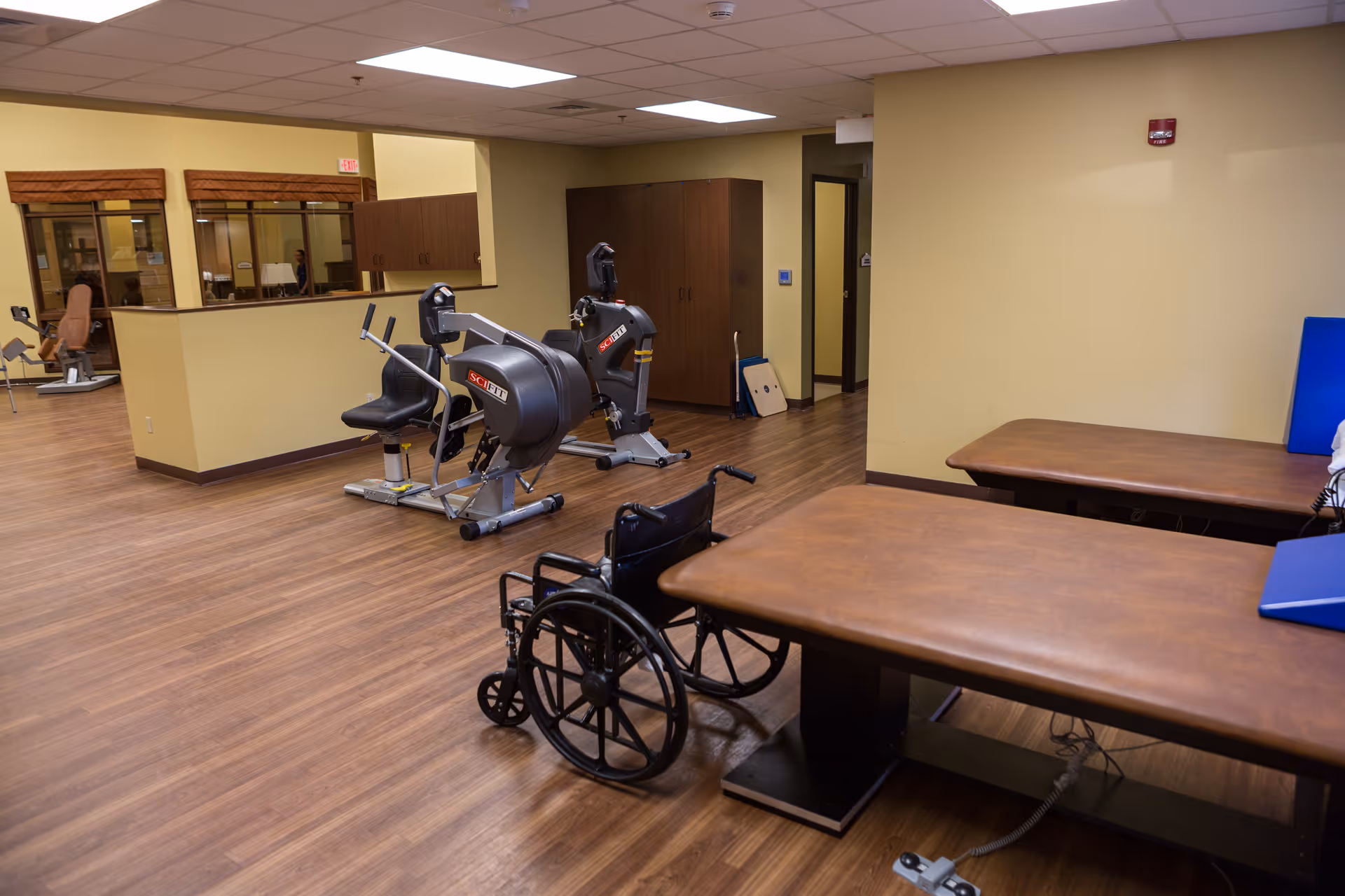 A rehabilitation or physical therapy room with exercise equipment, two brown therapy tables, and a black wheelchair. The room has wooden flooring, beige walls, and fluorescent ceiling lights.