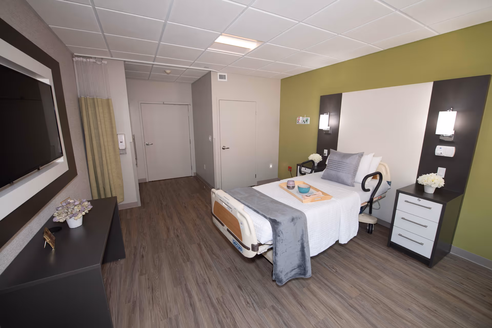 A clean and modern patient bedroom in a healthcare facility featuring a single hospital bed with white and gray bedding, a bedside table with a lamp and flowers, a flat-screen TV mounted on the wall, and wood-look flooring. The walls are painted in neutral tones with one green accent wall.