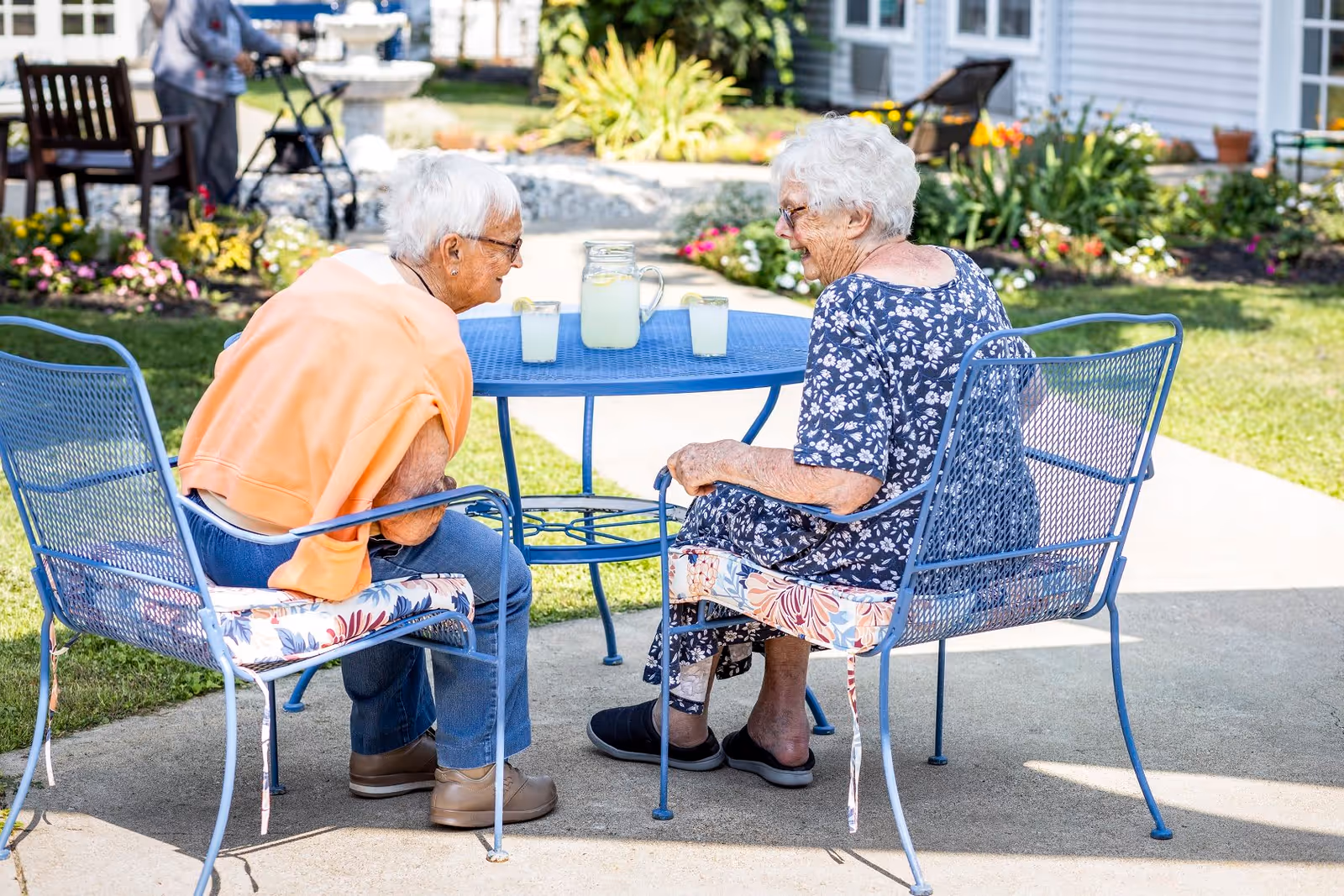 Two elderly women sitting outdoors at a blue metal table with matching chairs, enjoying lemonade on a sunny day in a garden area with flowers and greenery around.