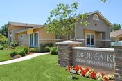 Exterior view of a single-story beige building with a well-maintained lawn, trees, and shrubs. In front of the building is a stone sign that reads 'Arbor Faire Senior Apartments' surrounded by red and white flowers.
