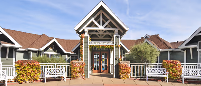 Front exterior view of Voralto at Belmont facility with a peaked roof entrance, white railings, benches, and plants with autumn-colored leaves on either side of the walkway.