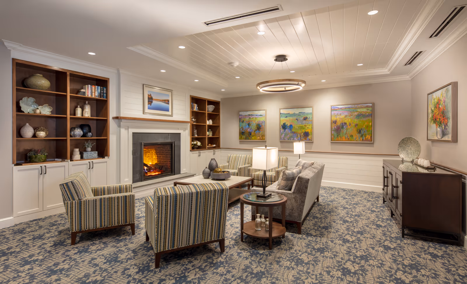 A cozy living room with a lit fireplace centered between two built-in wooden bookshelves. The room features a patterned carpet, a sofa, two striped armchairs, a coffee table, and side tables with lamps. The walls are decorated with colorful paintings and a floral artwork. The ceiling has recessed lighting and a modern circular chandelier.