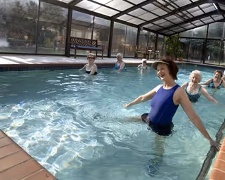 A group of elderly women participating in water exercise in an indoor swimming pool with a glass enclosure. The women are smiling and appear to be enjoying the activity.