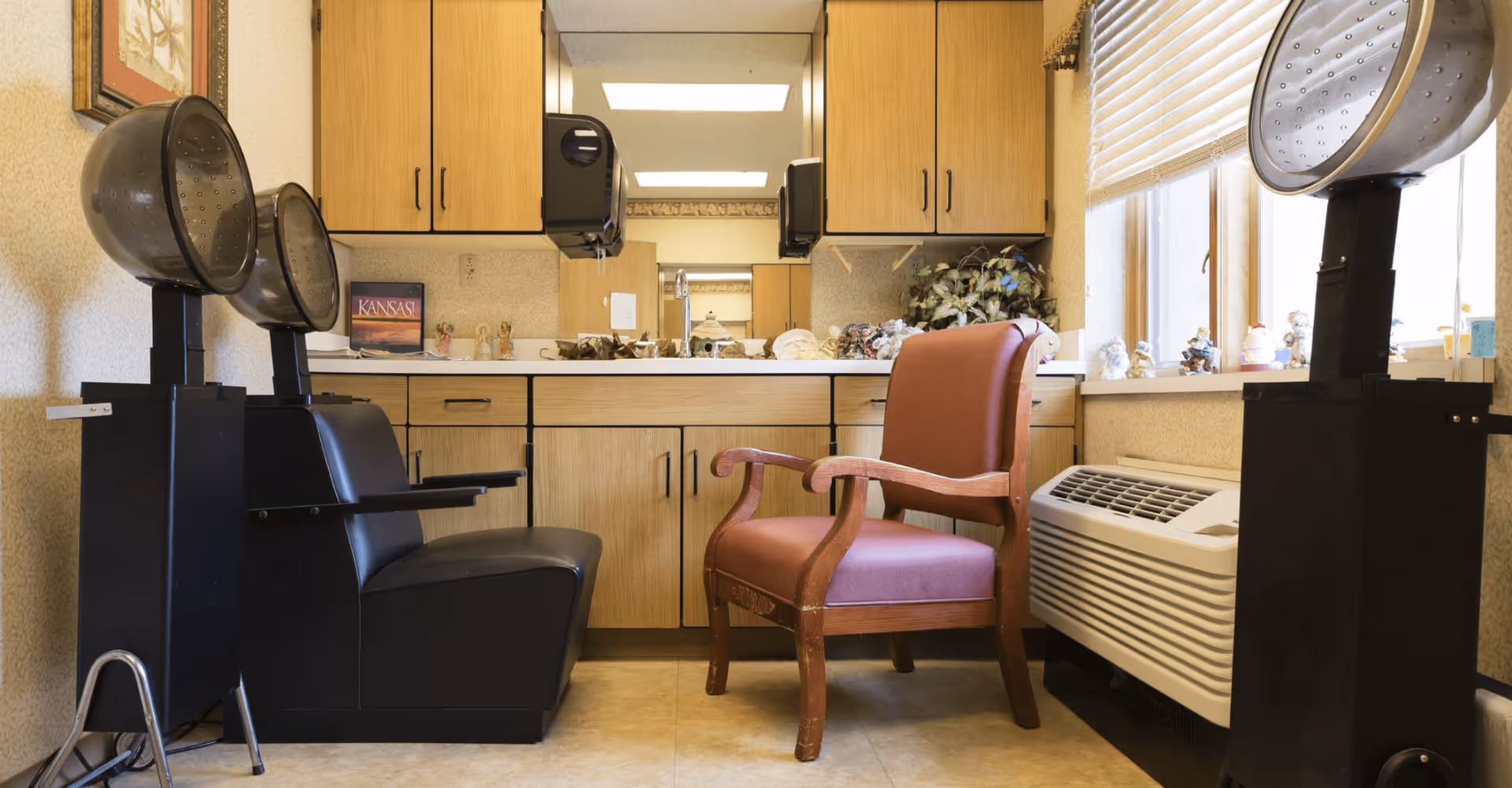 Interior view of a small salon area in an assisted living facility featuring two vintage hair dryer chairs, wooden cabinets with a countertop, a large mirror, and a window with blinds. Various small decorative items are placed on the countertop and windowsill.