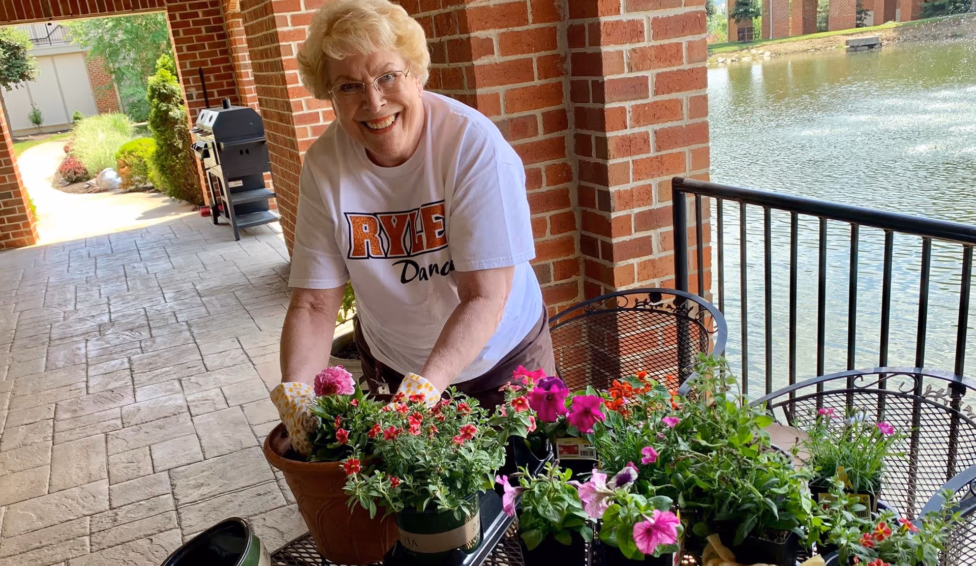 An elderly woman wearing glasses and gardening gloves is smiling while planting colorful flowers in pots on a patio next to a body of water with a fountain. The patio has brick pillars and metal chairs and tables.