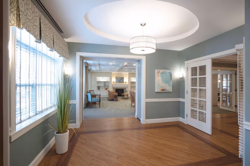 A bright and spacious interior hallway with wooden flooring and light blue walls. There is a large window with patterned blinds on the left, a potted plant beneath it, and a modern ceiling light fixture. The hallway opens into a living area with chairs, a fireplace, and artwork on the walls. To the right, there are white double doors with glass panels leading to another room.