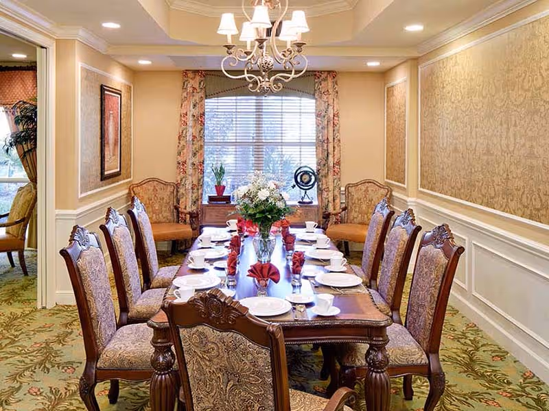 Formal dining room with a long set table, ornate upholstered chairs, chandelier, floral curtains, and place settings.
