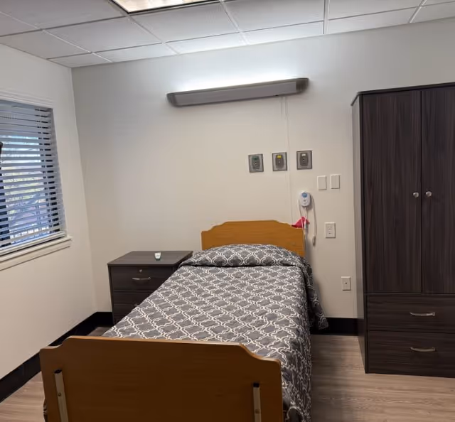 A single hospital-style bedroom with a bed, nightstand, and a tall wardrobe in a tidy room.