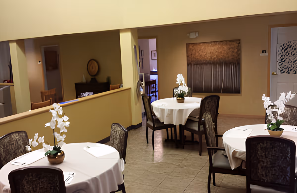 Interior view of a dining room with round tables covered with white tablecloths, each table decorated with a small potted white orchid. Several chairs with patterned upholstery surround the tables. The walls are painted beige and there is a large abstract painting on one wall. The room has a warm and inviting atmosphere.