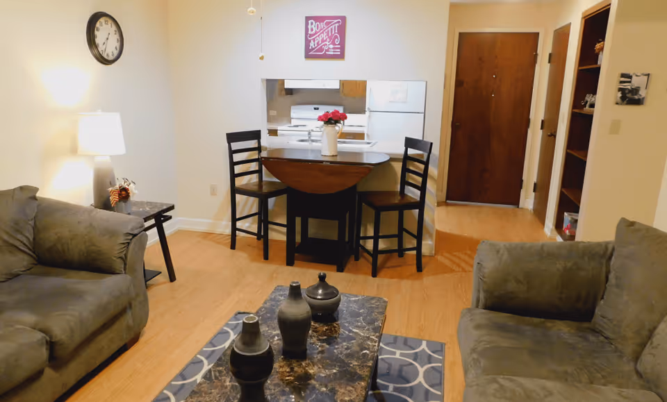 A cozy living room area with two gray sofas facing a marble coffee table with decorative vases. A small dining table with two chairs is positioned against a half wall that opens to a kitchen with a stove and refrigerator. A wall clock and a lamp on a side table are visible, along with a wooden door and a bookshelf in the background.