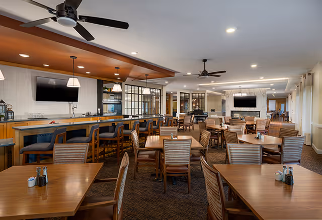A spacious dining room with multiple wooden tables and chairs arranged neatly. There is a bar area with high stools on the left side, pendant lights hanging above the bar, and ceiling fans. Two flat-screen TVs are mounted on the walls, one above the bar and another above a fireplace in the background. The room is well-lit with recessed lighting and large windows with curtains on the right side.