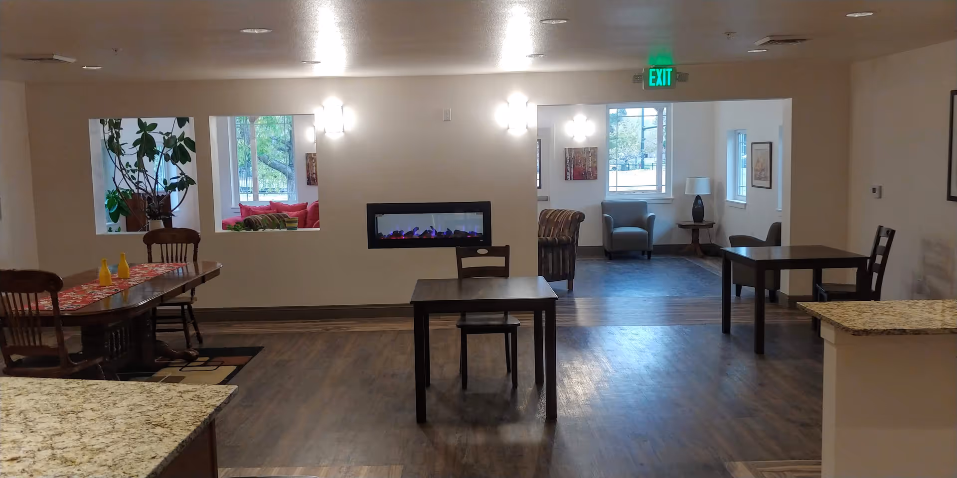 Interior view of a senior living facility common area with wooden tables and chairs, a modern electric fireplace mounted on a wall, and seating areas with armchairs near windows. The space has wood flooring, neutral-colored walls, and ceiling lights. An exit sign is visible above a doorway.