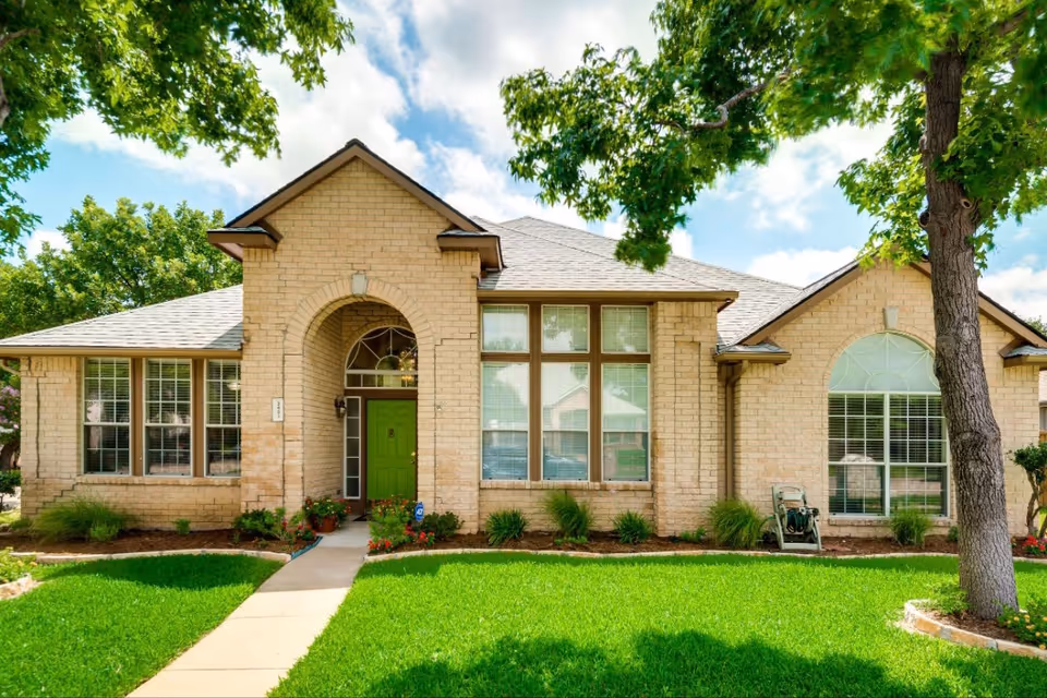 Front exterior view of a single-story brick house with a green front door, large windows, and a well-maintained lawn with trees and shrubs under a partly cloudy sky.