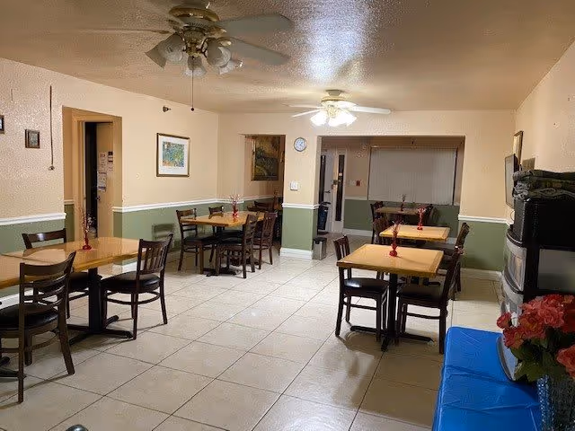 Communal dining room with multiple wooden tables and chairs, ceiling fans, tiled floor, and pale green wainscoting.