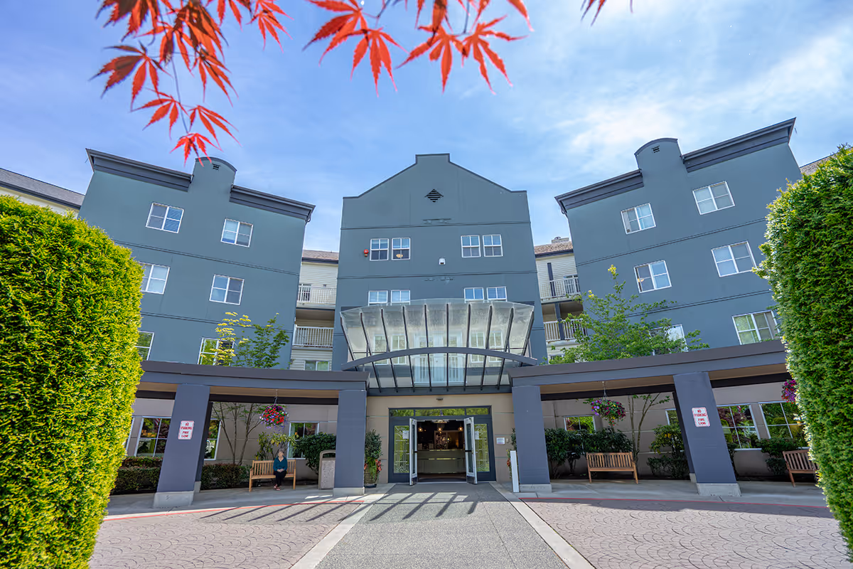Front entrance of a multi-story retirement building with a covered glass canopy, benches, and landscaped hedges.