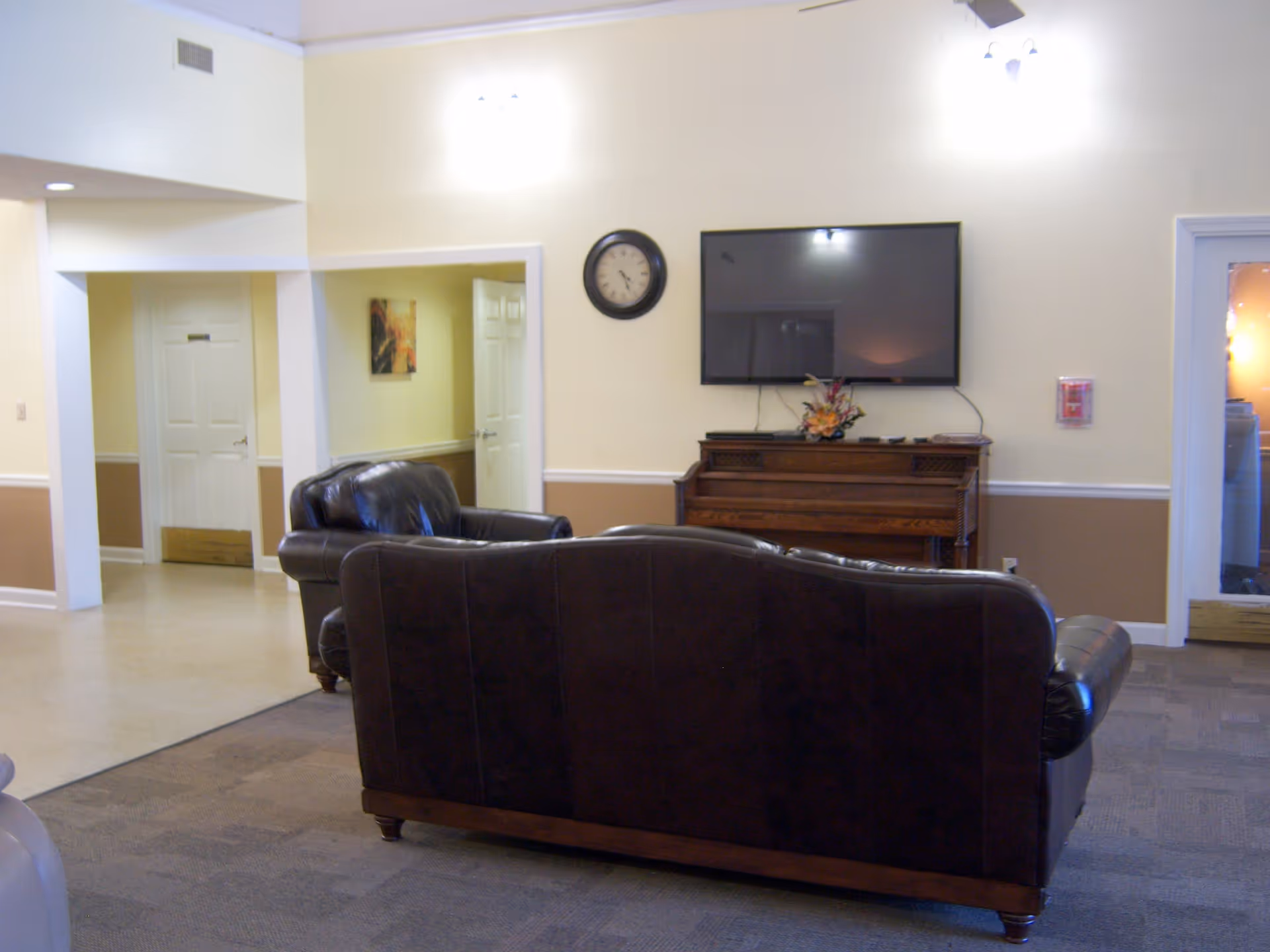 Interior view of a living room area with two dark brown leather sofas facing a wall-mounted flat screen TV above a wooden piano. The walls are painted beige with white trim, and there is a clock on the wall between two light fixtures. There are doorways leading to other rooms visible in the background.