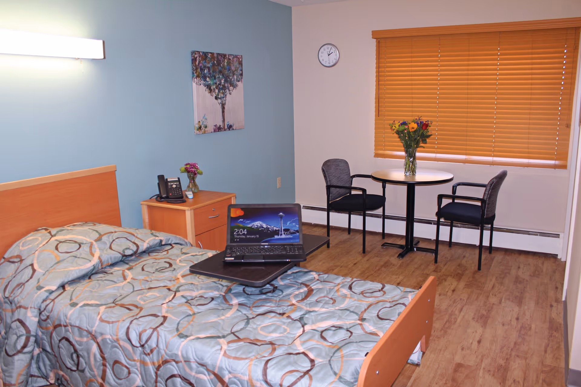 A senior living facility bedroom with a single bed covered in a patterned comforter. A laptop is placed on a tray table over the bed. Next to the bed is a wooden nightstand with a telephone and a small vase of flowers. On the wall above the nightstand is a colorful painting of a tree. Across from the bed is a small round table with two chairs and a vase of flowers. A window with wooden blinds is behind the table, and a clock is mounted on the wall above the table. The floor is wood laminate.