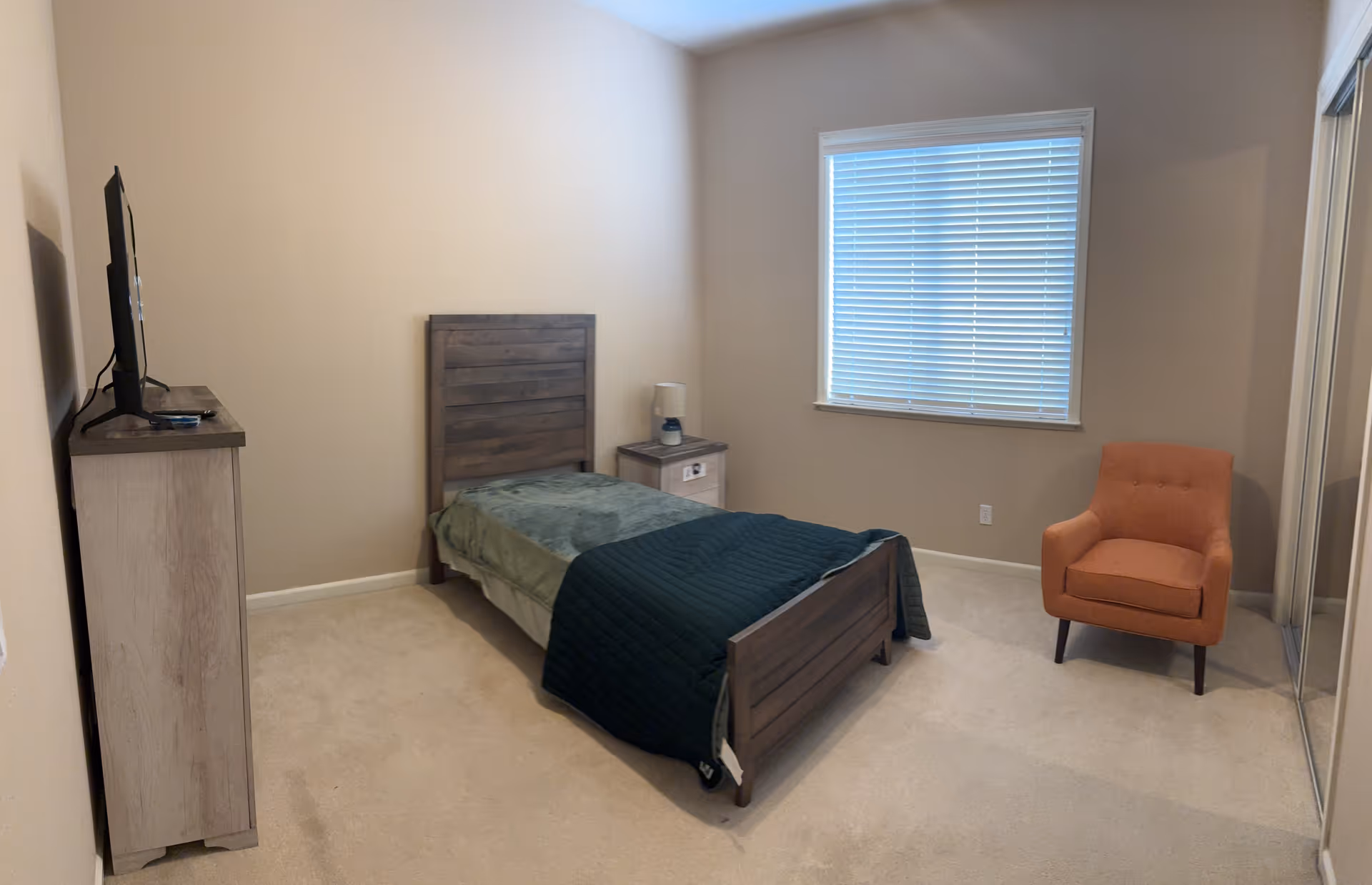 A simple bedroom with beige walls and carpet. The room features a single wooden bed with green bedding and a dark green blanket, a wooden nightstand with a lamp, a wooden dresser with a flat-screen TV on top, an orange upholstered armchair, and a window with closed blinds. There is also a mirrored closet door on the right side.