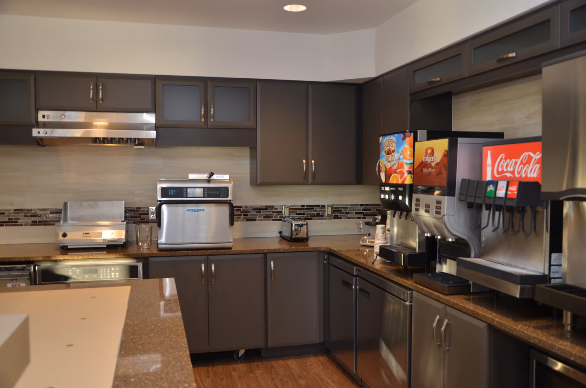 Interior view of a kitchen area with dark cabinets, a countertop with various appliances including a toaster oven, a microwave, a coffee dispenser, and a Coca-Cola soda fountain machine.