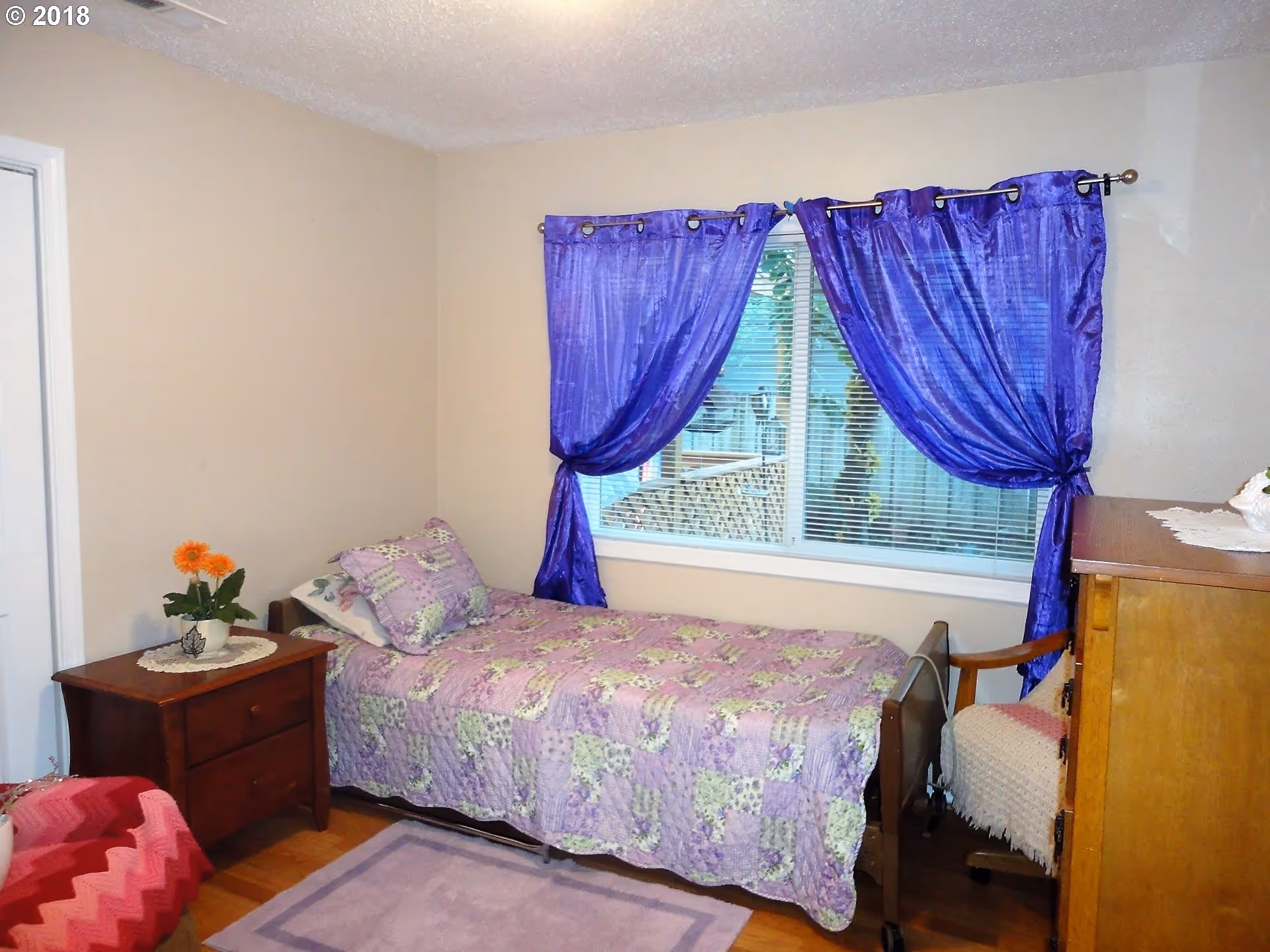 A small bedroom with a single bed covered in a purple and green patchwork quilt. The bed is positioned against a wall with a window that has purple curtains tied back. Next to the bed is a wooden nightstand with a small potted plant with orange flowers. There is a wooden dresser and a chair with a white knitted cover in the corner. The walls are beige and the floor is wooden with a small purple rug.
