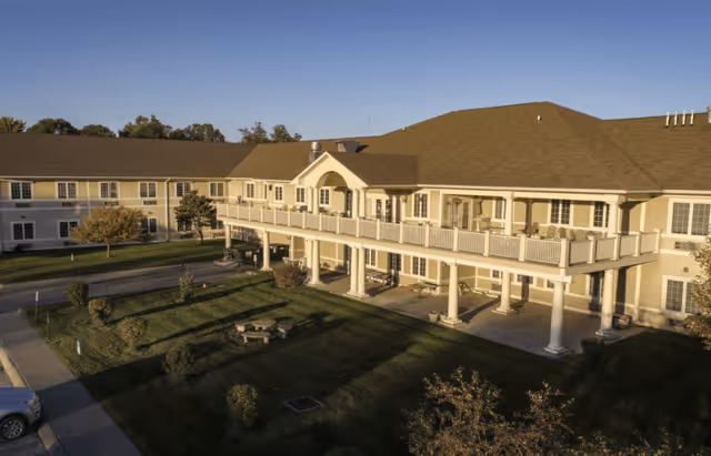 Exterior view of a two-story assisted living facility building with beige siding and a brown roof. The building features a large balcony with white railings supported by columns. There is a well-maintained lawn with small bushes and a picnic table in front of the building. The sky is clear and blue.