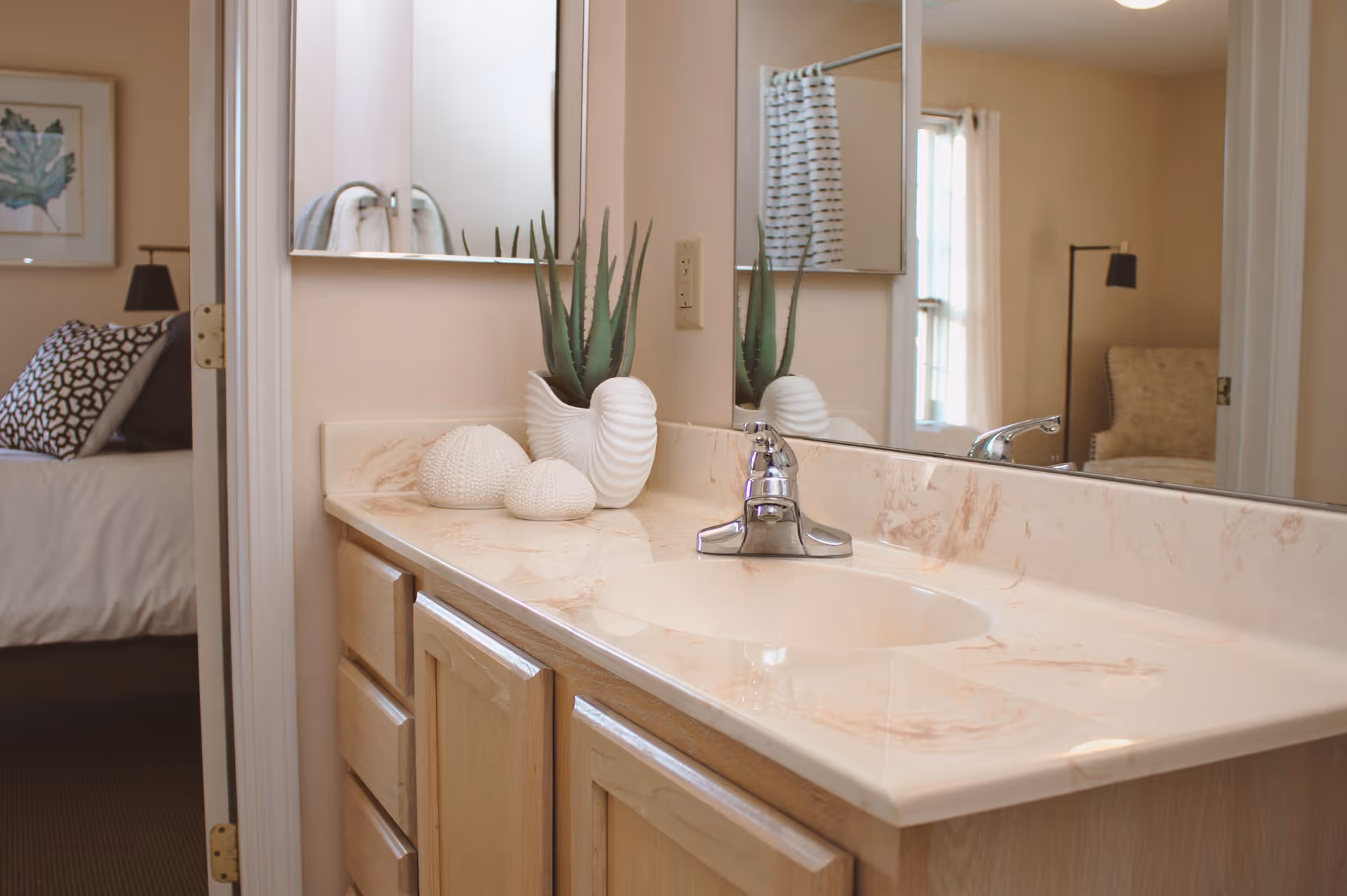 A bathroom vanity with a beige marble countertop, a built-in sink, and a chrome faucet. On the countertop, there is a white decorative vase with a green succulent plant and two white textured decorative objects. A large mirror is mounted on the wall above the vanity. In the background, a bedroom with a bed, pillows, a framed picture, and a floor lamp is partially visible through an open door.