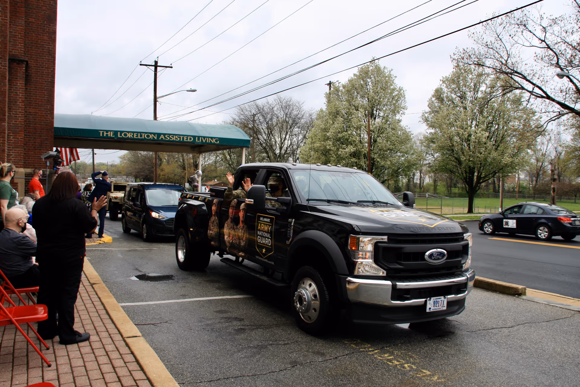 A black Ford pickup truck with Army National Guard graphics drives past the entrance of The Lorelton Assisted Living facility. Several people stand and sit on the sidewalk, waving at the passing vehicles. Trees and a road are visible in the background under a cloudy sky.