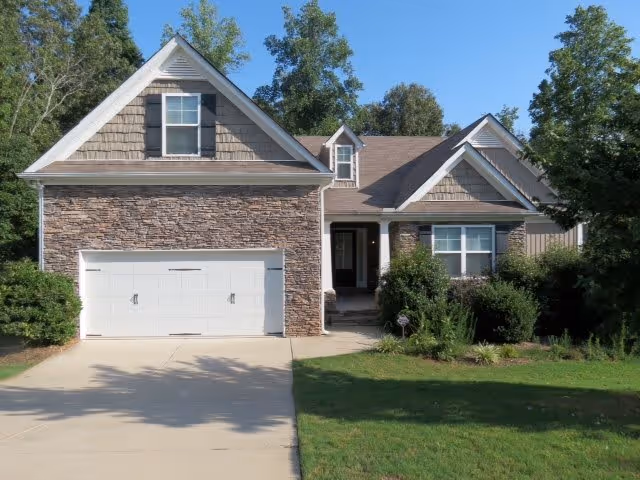 Front exterior view of a single-story house with a stone facade, a two-car garage, and a driveway. The house is surrounded by green grass, bushes, and trees under a clear blue sky.
