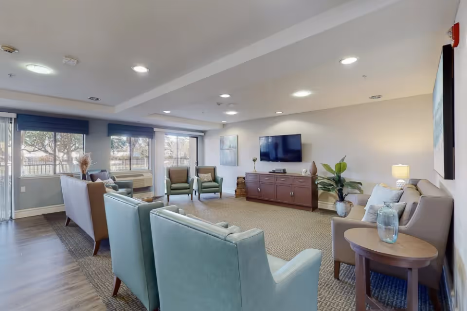 A spacious and well-lit living room with multiple armchairs and sofas arranged around a large carpet. There is a flat-screen TV mounted on the wall above a wooden cabinet, and large windows with blue blinds letting in natural light. The room is decorated with plants, lamps, and artwork on the walls.