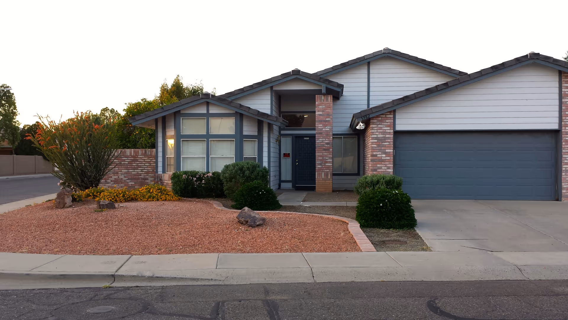 Front exterior view of a single-story house with gray siding and brick accents, a dark blue front door, a two-car garage, and a landscaped front yard with bushes, rocks, and gravel.