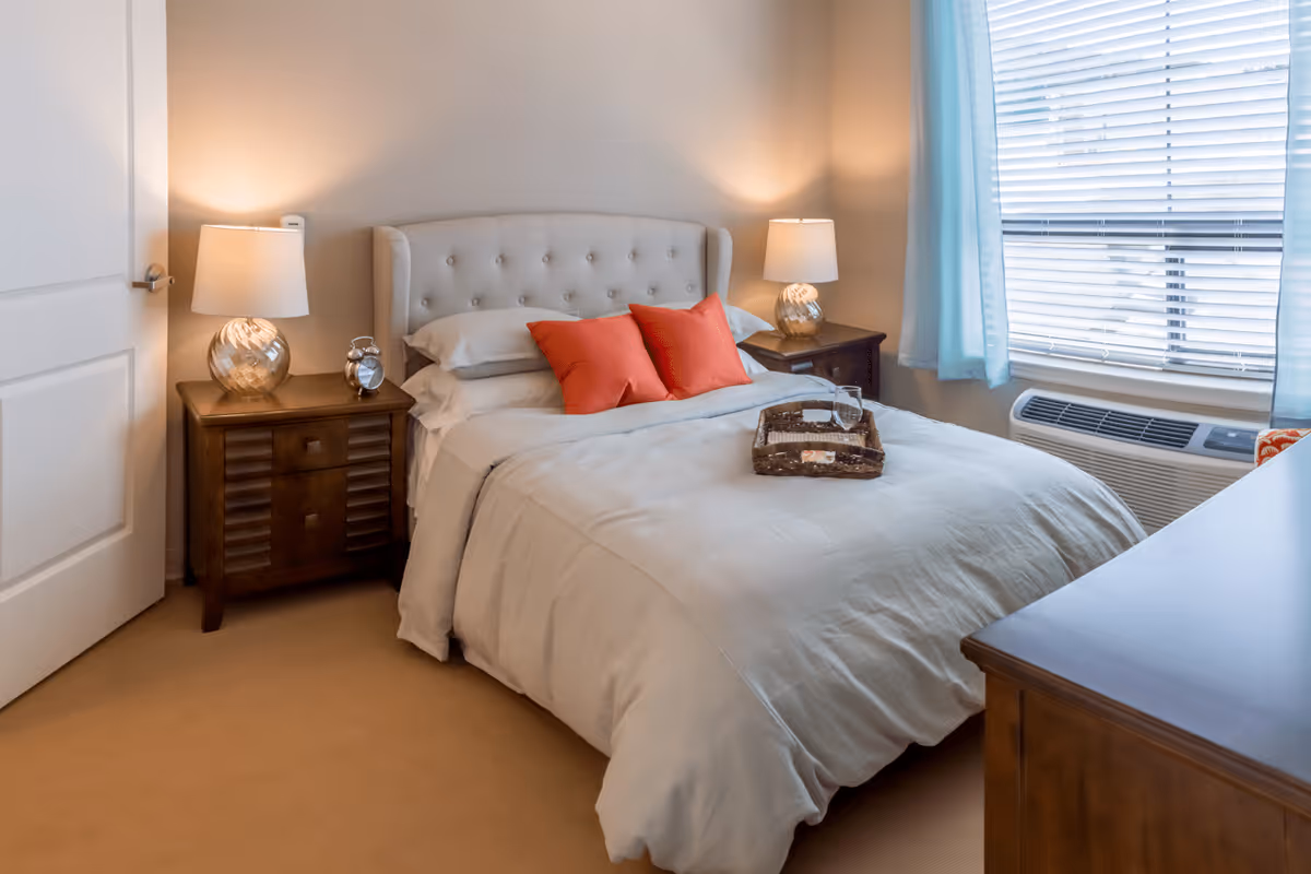 A cozy bedroom with a beige upholstered headboard bed, white bedding, and two red accent pillows. There are two wooden nightstands on either side of the bed, each with a lamp. A window with blue curtains and blinds is on the right side, and a wooden dresser is partially visible in the foreground.