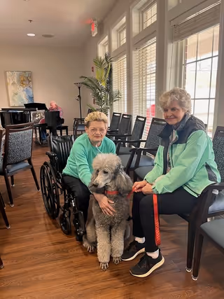 Two elderly women sitting in a well-lit room with large windows. One woman is in a wheelchair petting a large gray poodle, while the other woman sits on a chair holding the dog's leash. In the background, there is a man playing a grand piano and several empty chairs arranged along the wall.